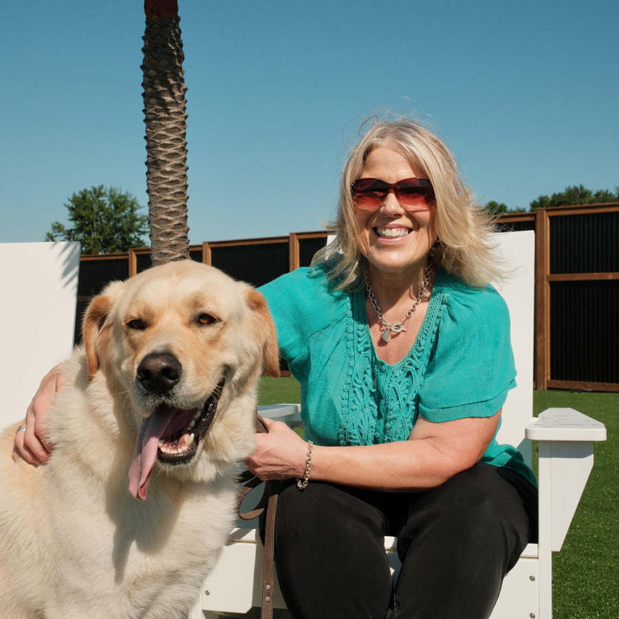 A woman wearing a turquoise short-sleeve top and black pants sits on a white outdoor chair next to a golden retriever dog, with a palm tree and blue sky in the background.
